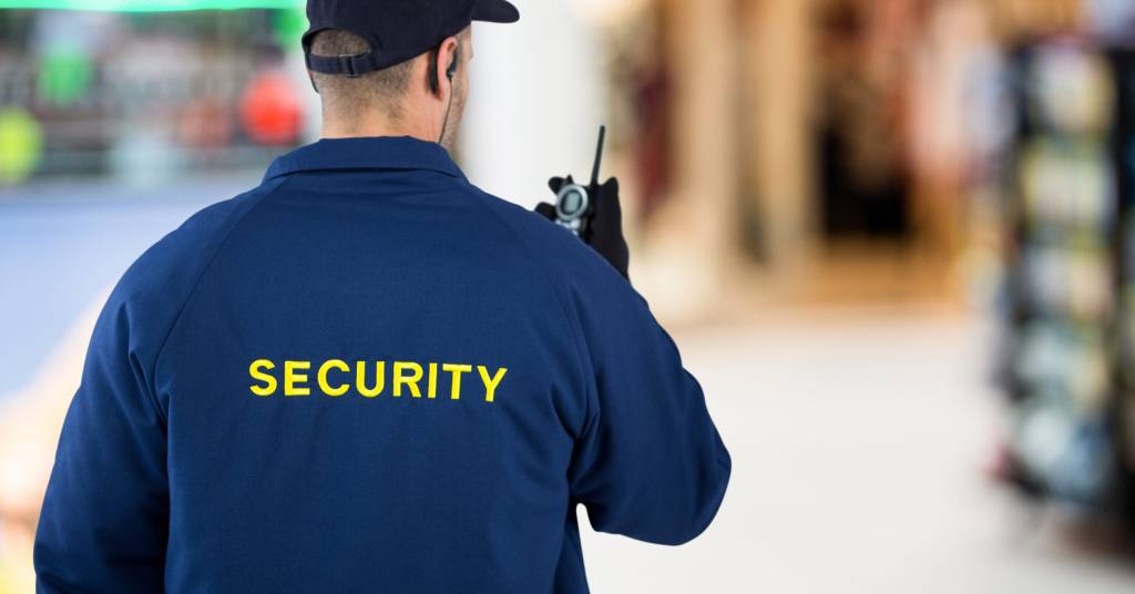 A security guard in a blue jacket with "SECURITY" in yellow on the back holds a walkie-talkie. The background is a blurred indoor setting.